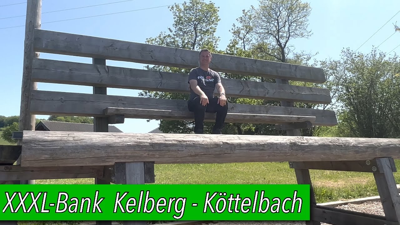Mann sitzt auf einer riesigen Holzbank im Freien vor Bäumen und blauem Himmel.